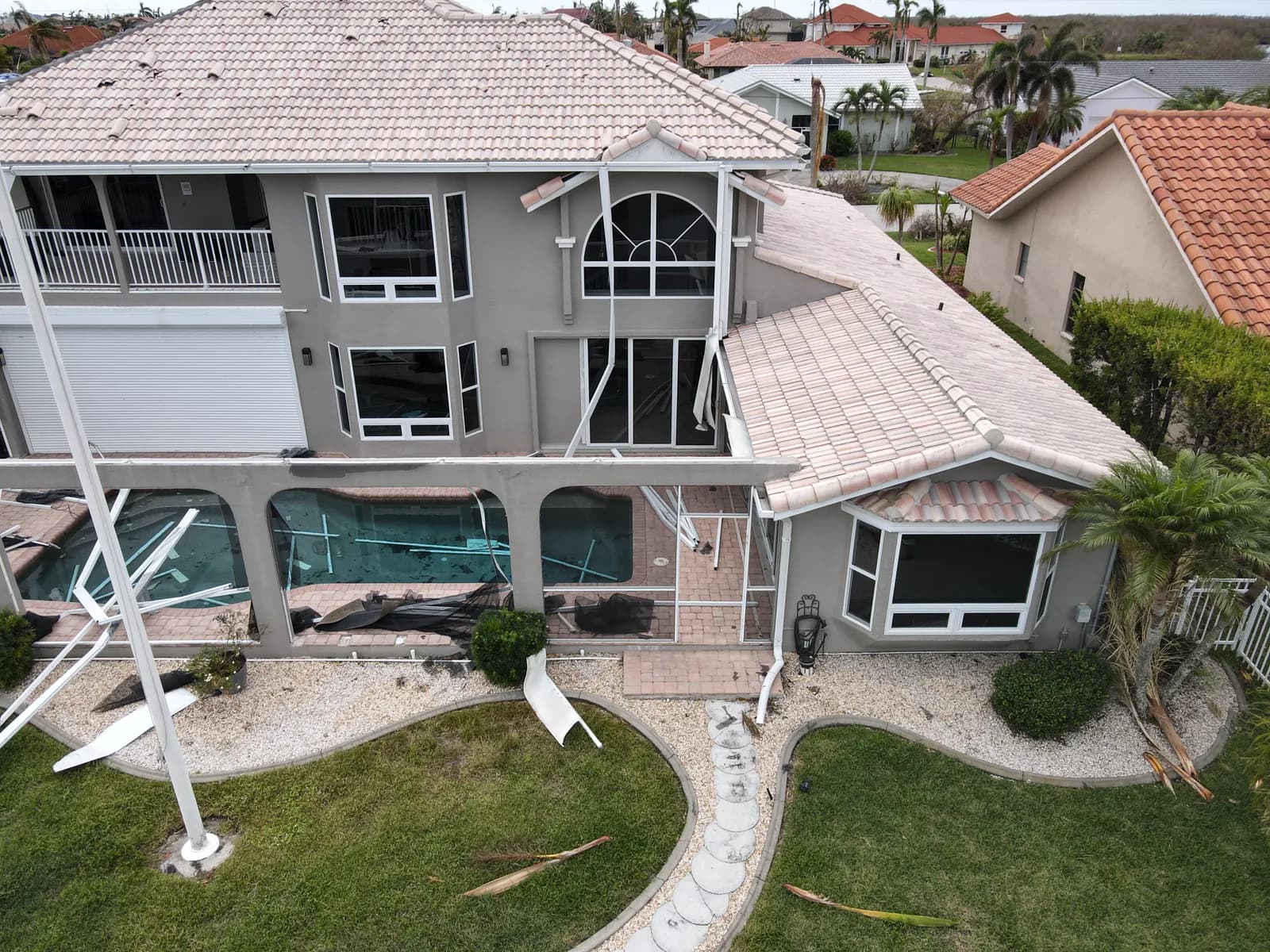 Aerial drone view of a two-story stucco home with a concrete tile roof after a hurricane, showing a torn pool screen enclosure and a bent flagpole in the yard