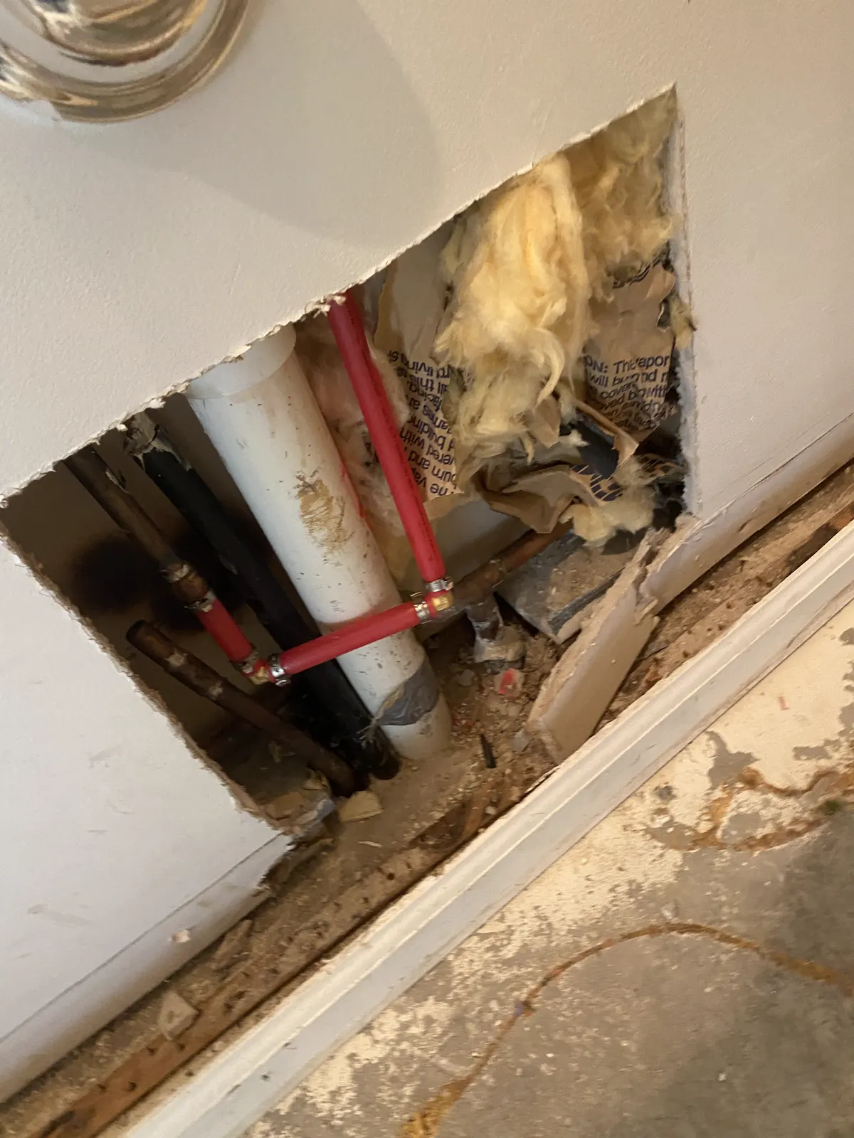 Drywall cut open at the base of a wall to expose a PVC drain, red PEX supply lines and wet insulation above a stained subfloor