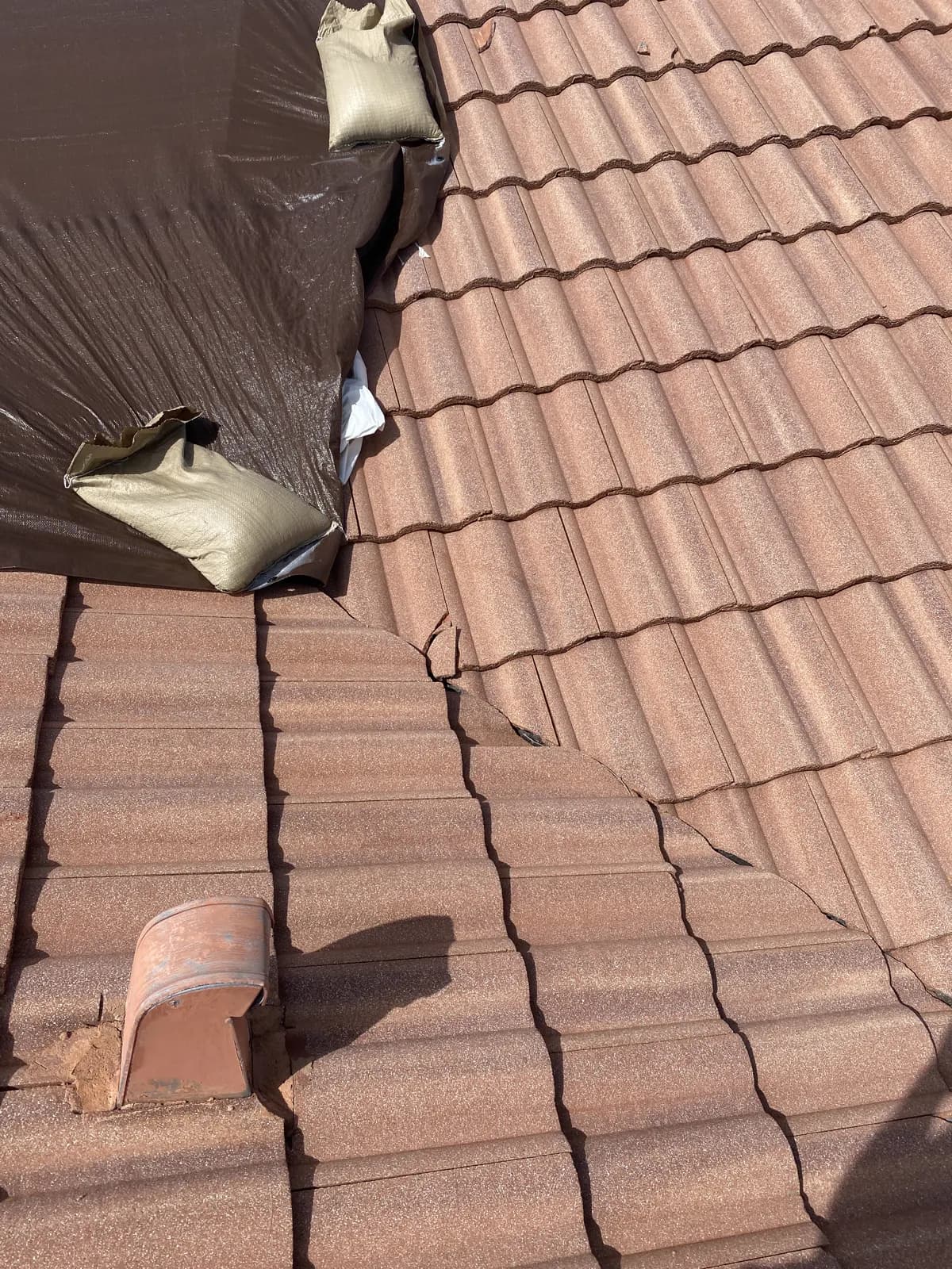 Barrel tile roof with a dark emergency tarp held down by sandbags beside displaced and broken tiles
